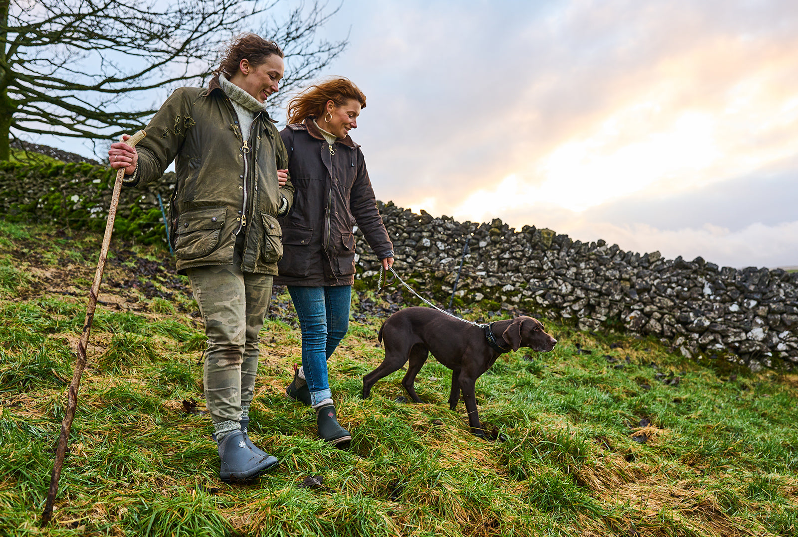 Zwei Frauen in Muck Boots Original Ankleboots gehen mit einem Hund auf einem Feld spazieren, im Hintergrund ist eine Steinmauer