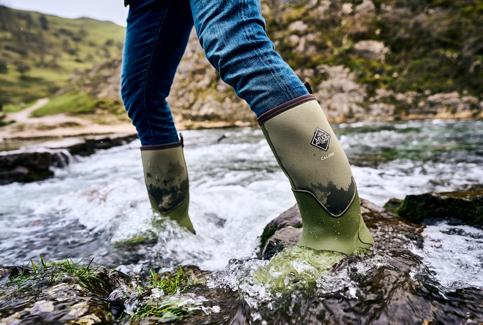 Person, die durch einen felsisgen Bach geht und ein Paar Muck Boots Calder Gummistiefel trägt