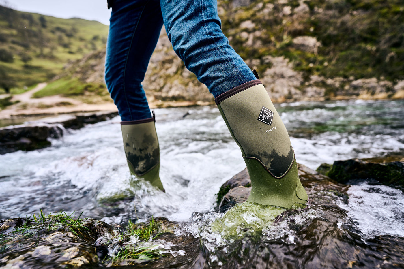 Person, die mit Muck Boots Calder Gummistiefeln einen Bach mit Felsen überquert