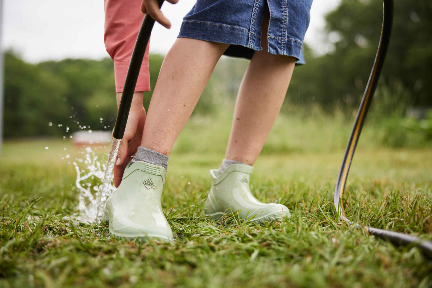 Nahaufnahme einer Person, die ein Paar grüne Muck Boot Muckster Lite Stiefel trägt, die Unterseite ihres Absatzes mit einem Schlauch abwäscht und den oberen Teil des Absatzes festhält