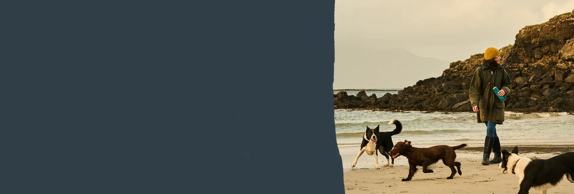 Eine Frau in Muck Boots Gummistiefeln und einer gelben Dockermütze am Strand mit drei Hunden.