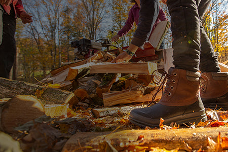 Eine Person sortiert gehacktes Holz in einem Paar Muck Boot Duckboots und Herbstblätter liegen auf dem Boden.