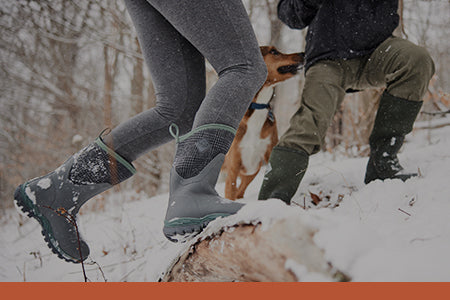 Zwei Personen in Muck Boot Gummistiefeln spielen im Schnee mit einem Hund