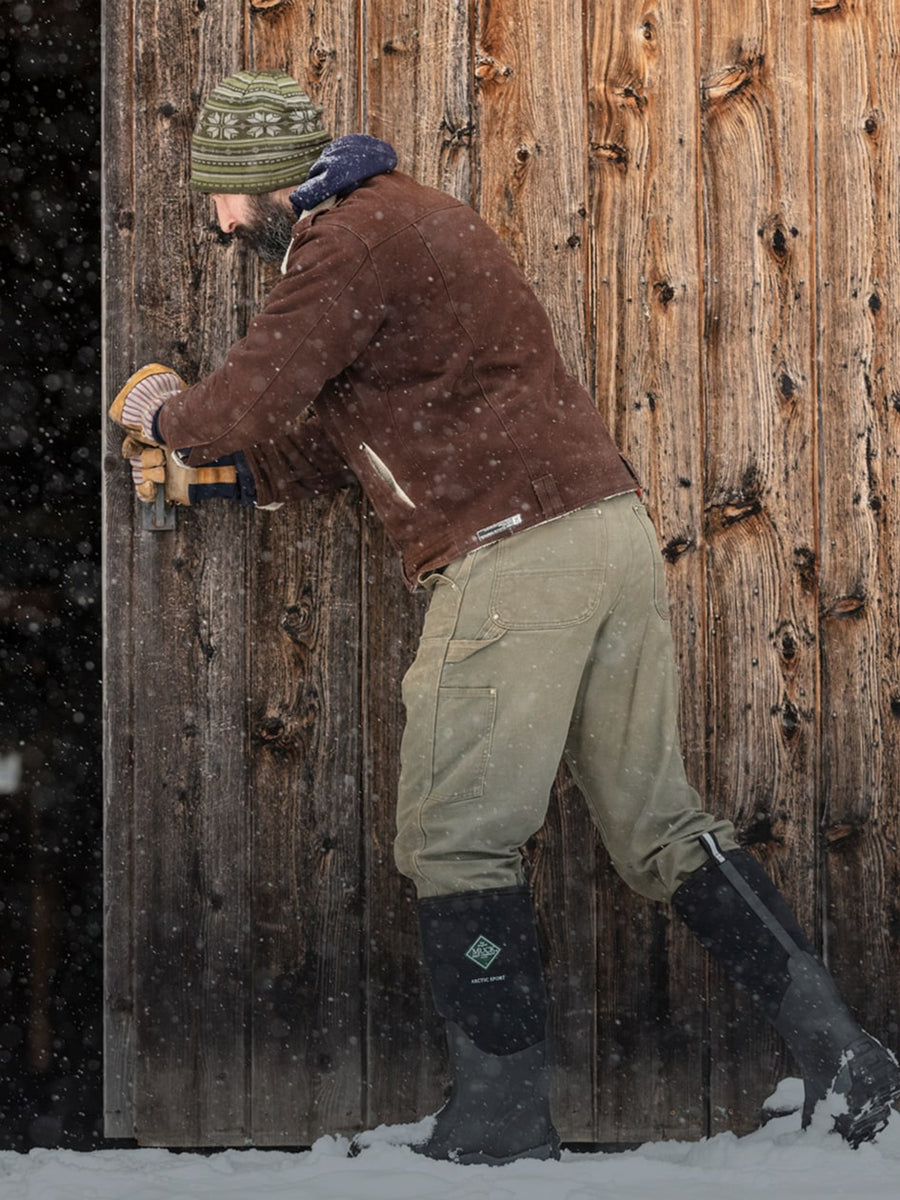 Nahaufnahme einer Person, die im Schnee vor einem offenen Scheunentor aus Holz ein Paar Muck Boots Arctic Sport Gummistiefel trägt