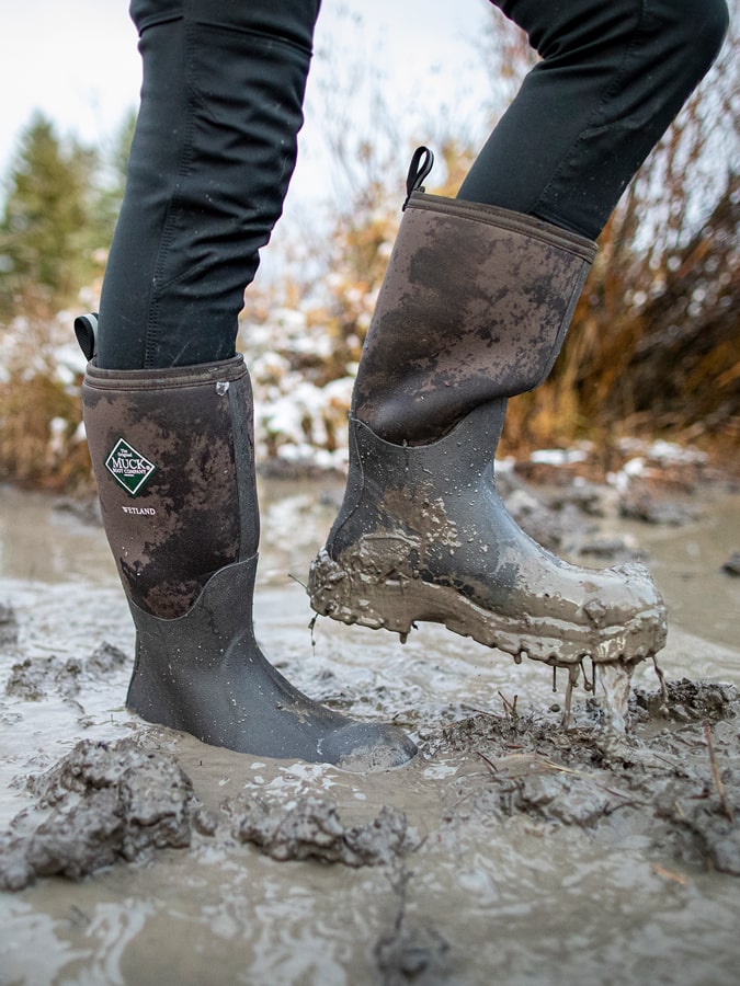 Nahaufnahme einer Person, die ein Paar mit Schlamm überzogene Muck Boots Wetland Gummistiefel trägt.