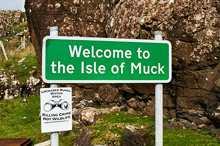 Ein grünes Schild mit der Aufschrift “Welcome to the Isle of Muck” vor Felsen und Geröll