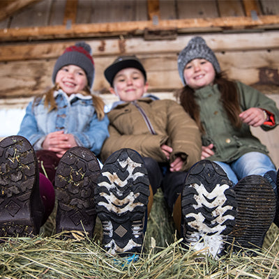 Kinder sitzen auf einem Heuhaufen in einer Scheune und tragen Muck Boot Gummistiefel