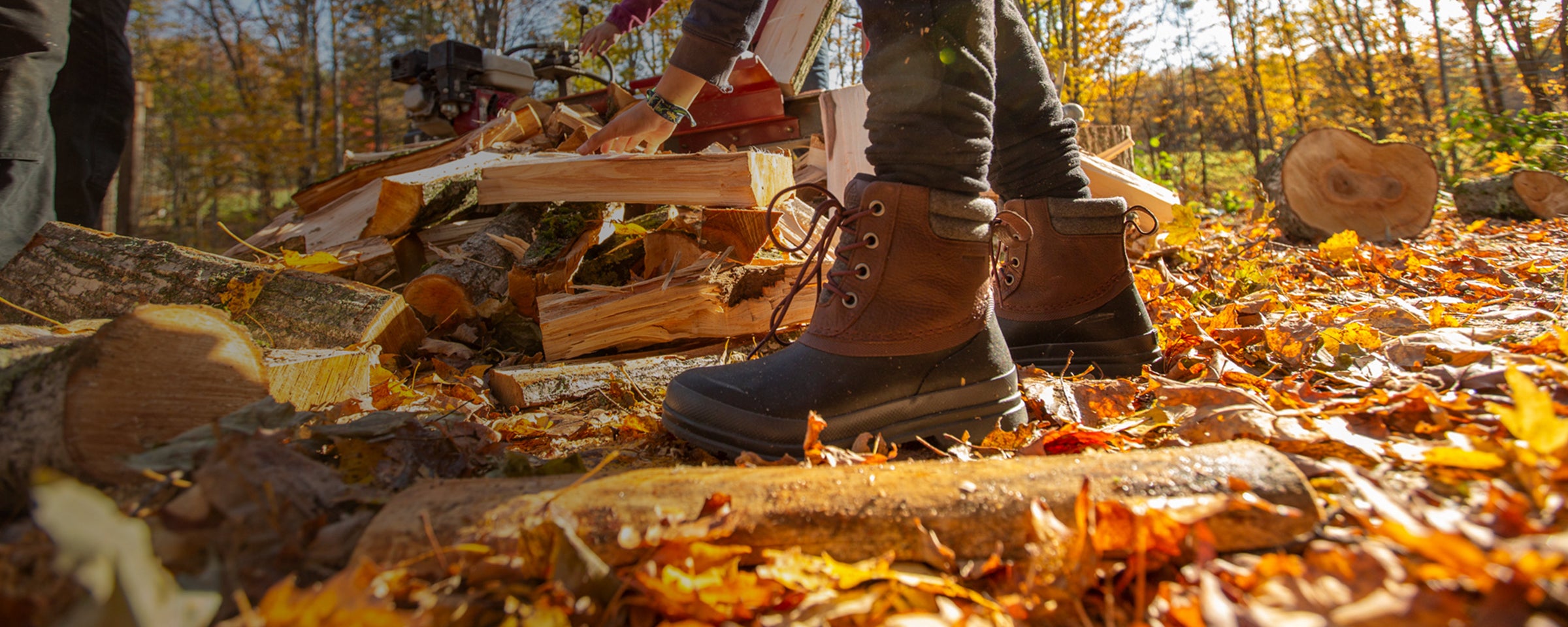 Eine Person sortiert gehacktes Holz in einem Paar Muck Boot Duckboots und Herbstblätter liegen auf dem Boden.