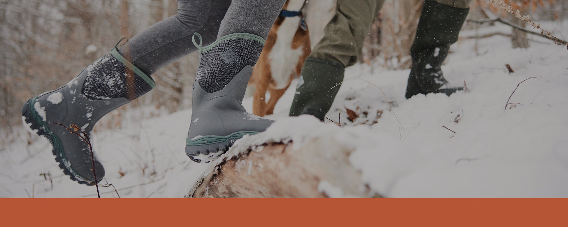 Zwei Personen in Muck Boot Gummistiefeln spielen im Schnee mit einem Hund