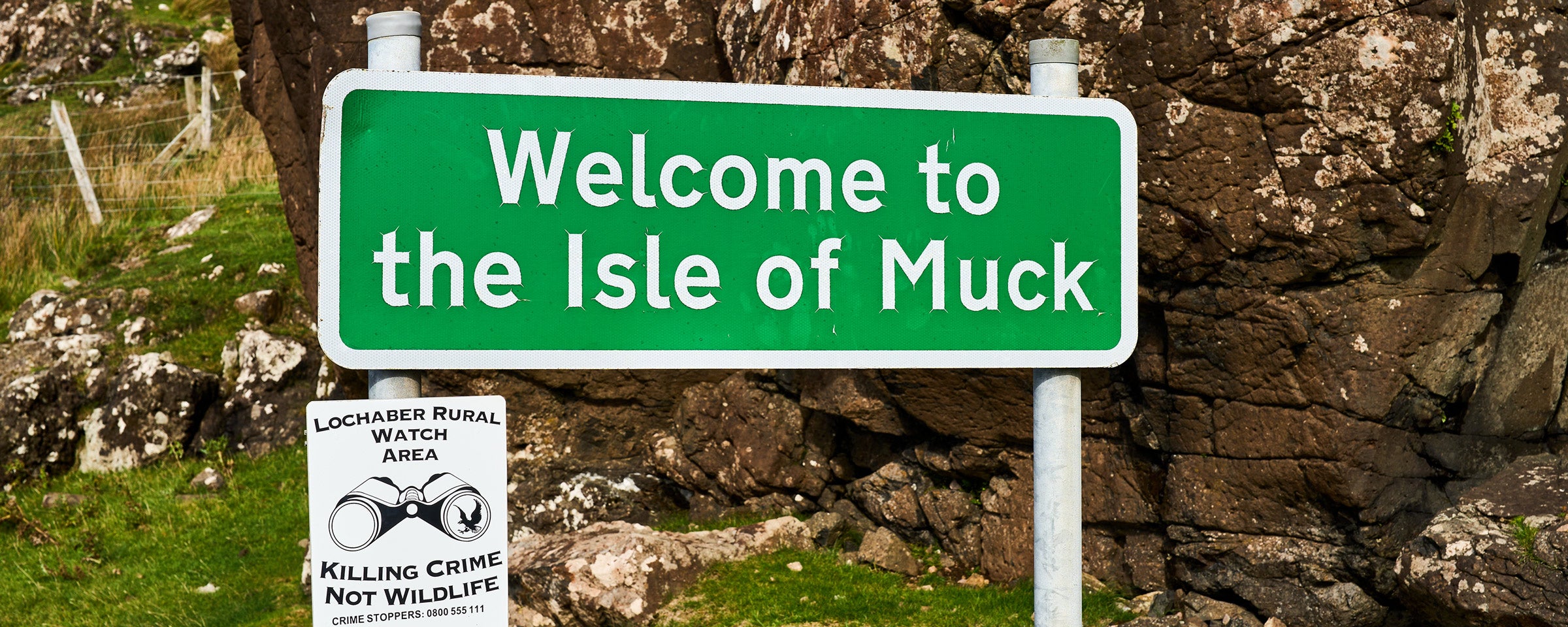 Ein grünes Schild mit der Aufschrift “Welcome to the Isle of Muck” vor Felsen und Geröll