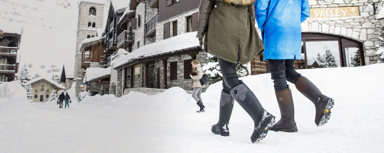 Ein Mann und eine Frau spazieren auf einer schneebedeckten Straße entlang, im Hintergrund sind Berge zu sehen. Sie tragen warme Kleidung und Muck Boots Gummistiefel.