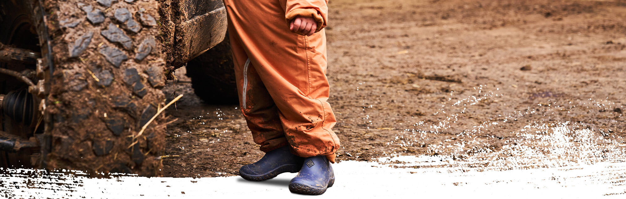 Ein Kleinkind in orangefarbener Latzhose und blauen Muck Boots Mini Mucks Stiefeln steht neben einem großen Reifen auf schlammigem Boden.
