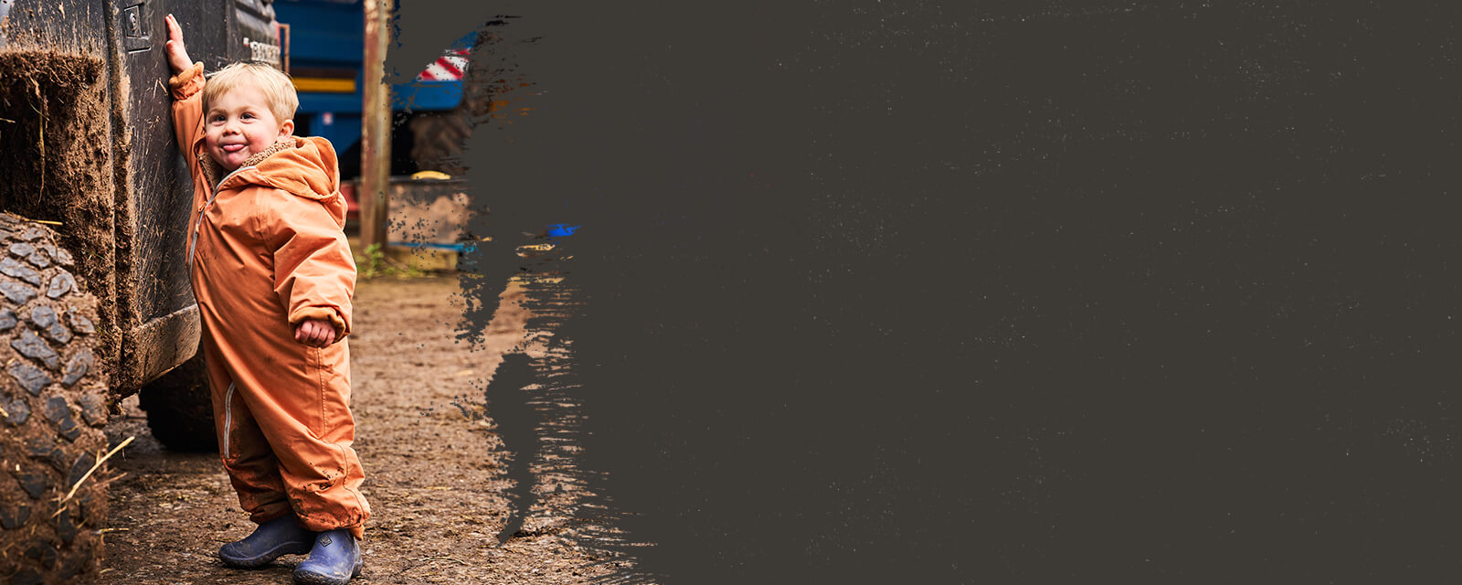 Ein Kind, das in einem blauen Paar Muck Boots Kinder Muckster Stiefel und in einem orangen Spielanzug neben einem großen Reifen vor einem dunklen Hintergrund steht