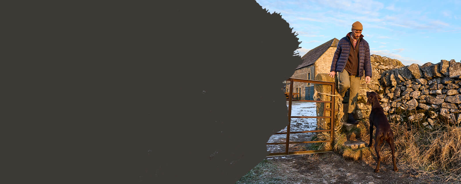 Eine Person mit einem Hund, die in der Nähe eines Metalltors vor einer malerischen Winterkulisse auf einer Steinmauer steht.