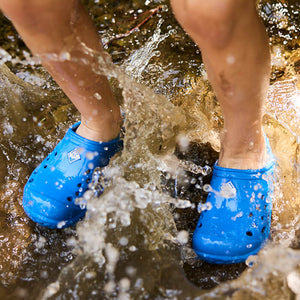 Nahaufnahme eines Kindes, das im Wasser planscht und ein Paar blaue Muck Boots Muckster Lite Clogs trägt.
