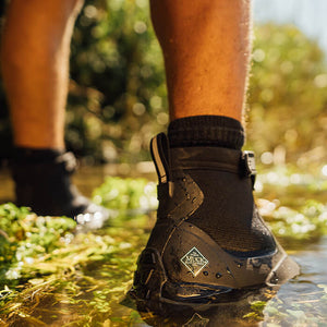 Nahaufnahme eines Mannes, der im Wasser steht und ein Paar schwarze Muck Boots Apex Stiefel trägt.
