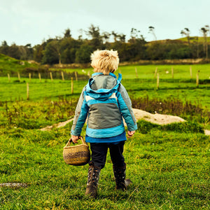 Ein Kind in Muck Boots und einer blau-grauen Jacke. das mit einem Korb in der Hand über eine Wiese geht.