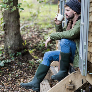 Ein Mann, der in einem grünen Paar Muck Boots Arctic Outpost Gummistiefel und einer grünen Jacke auf einer Holzstufe im Freien sitzt, und einen weißen Becher hält.