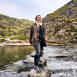 Eine Frau in einem Paar Gummistiefel von Muck Boots, die auf einem Felsen in einem Fluss steht, mit Bergen im Hintergrund.