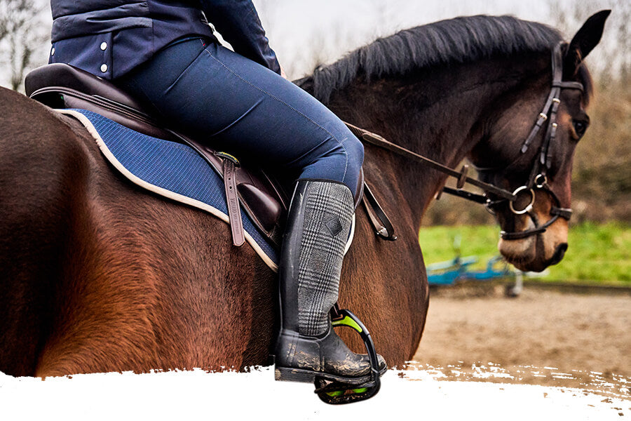 Eine Frau in einem Paar Muck Boots Damen Derby Stiefeln, die auf einem braunen Pferd auf einem Feldweg vor unscharfem Hintergrund reitet