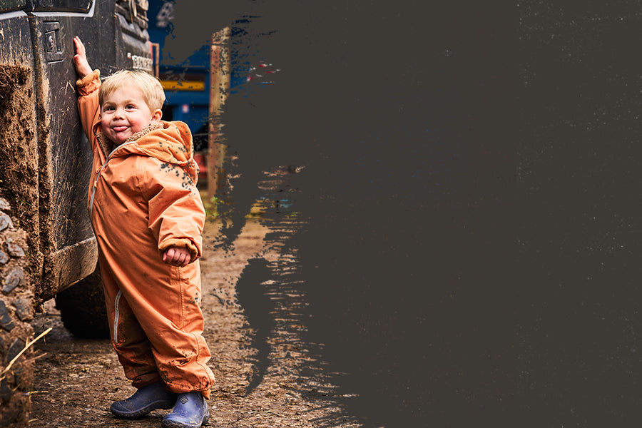 Ein Kind, das in einem blauen Paar Muck Boots Kinder Muckster Stiefel und in einem orangen Spielanzug neben einem großen Reifen vor einem dunklen Hintergrund steht