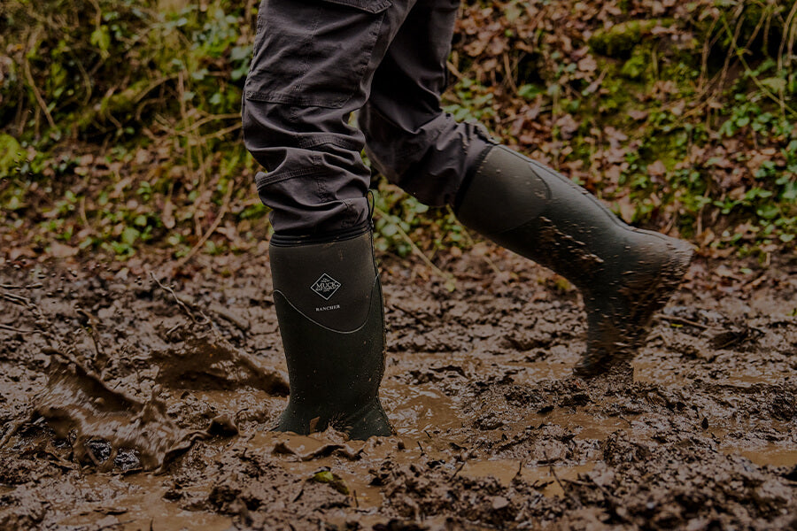 Eine Person, die in einem grünem Paar Muck Boots Rancher Gummistiefel durch ein schlammiges Feld läuft