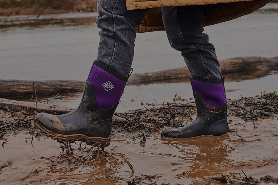 Eine Person läuft in einem Paar Muck Boots Damen Arctic Sport II Stiefel in Schwarz/Acai durch eine schlammige Pfütze.