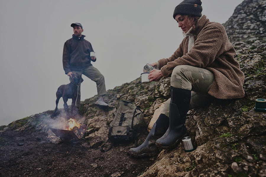 A man and woman on a rocky hillside, the woman pouring a hot drink with a fire burning and the man standing next to a dog. They are both wearing Muck Boots winter wellingtons.