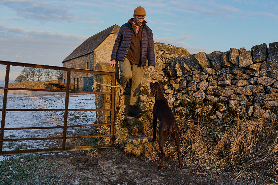 Eine Person mit einem Hund, die in der Nähe eines Metalltors vor einer malerischen Winterkulisse auf einer Steinmauer steht.