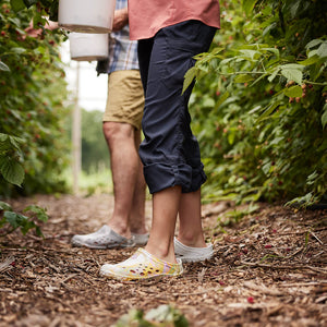 Eine Frau in Shorts und Muck Boots Apex Stiefeln, die Holzscheite in eine Feuerschale außerhalb eines Zeltes im Wald legt.