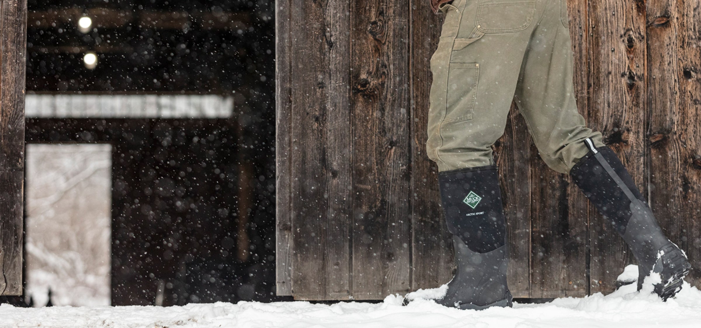 Nahaufnahme einer Person, die im Schnee vor einem offenen Scheunentor aus Holz ein Paar Muck Boots Arctic Sport Gummistiefel trägt