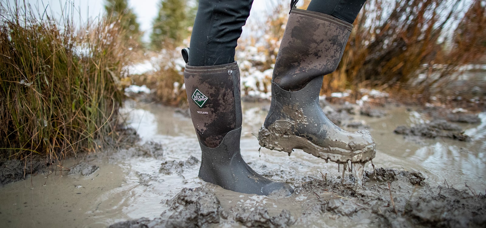 Nahaufnahme einer Person, die ein Paar mit Schlamm überzogene Muck Boots Wetland Gummistiefel trägt.