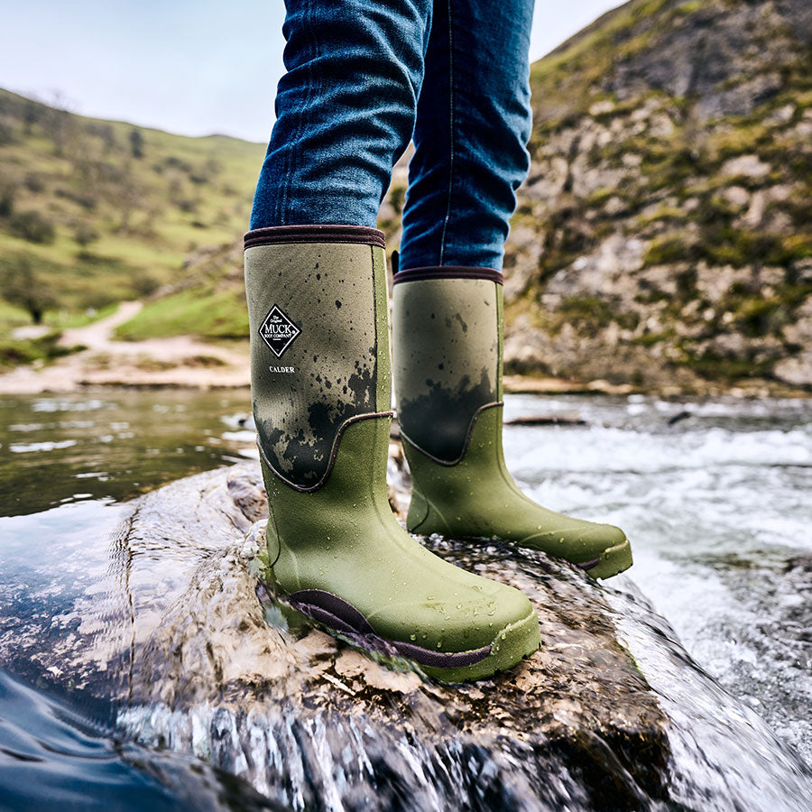 Nahaufnahme eines Mannes, der in einem grünen Paar Muck Boot Calder Gummistiefel auf einem Felsen in einem schnell fließenden Fluss steht.