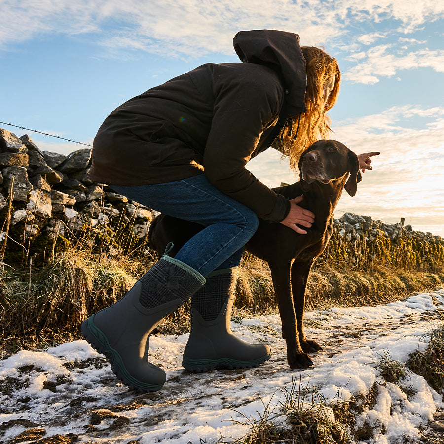 Eine Frau mit einem Hund bückt sich und zeigt auf einem verschneiten Weg nach vorne. Sie trägt eine Jacke, Jeans und ein Paar Muck Boots Stiefel.