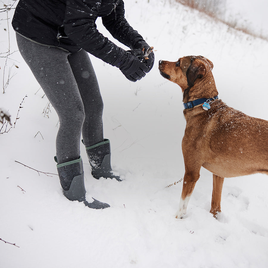 Eine Person trägt ein Paar Muck Boots Stiefel im Schnee und zeigt einem Hund einen Stock