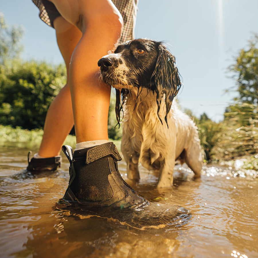 Eine Frau, die ein Paar Muck Boots trägt und mit einem Hund in einem Bach steht