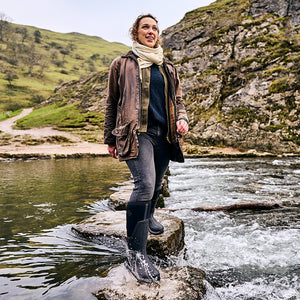 Eine Frau, die einen Schal und eine Wachsjacke trägt, steht in einem Tal in einem Paar Muck Boot Gummistiefel auf Felsen in einem Fluss