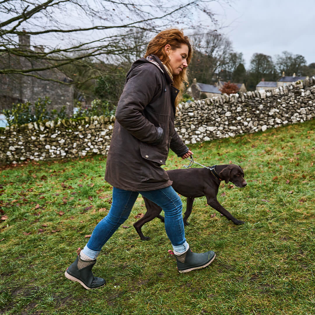 Frau in Stiefeln von Muck Boots, die mit einem schwarzen Hund auf einer Wiese mit Bäumen und einer Steinmauer im Hintergrund spazieren geht.