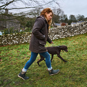 Frau in Stiefeln von Muck Boots, die mit einem schwarzen Hund auf einer Wiese mit Bäumen und einer Steinmauer im Hintergrund spazieren geht.