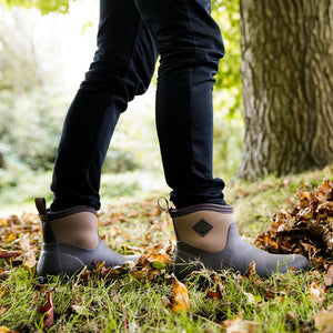 Eine Person in einem Paar Muck Boots Muckster II Stiefel vor einem Hintergrund mit einem Baumstamm und Gras.