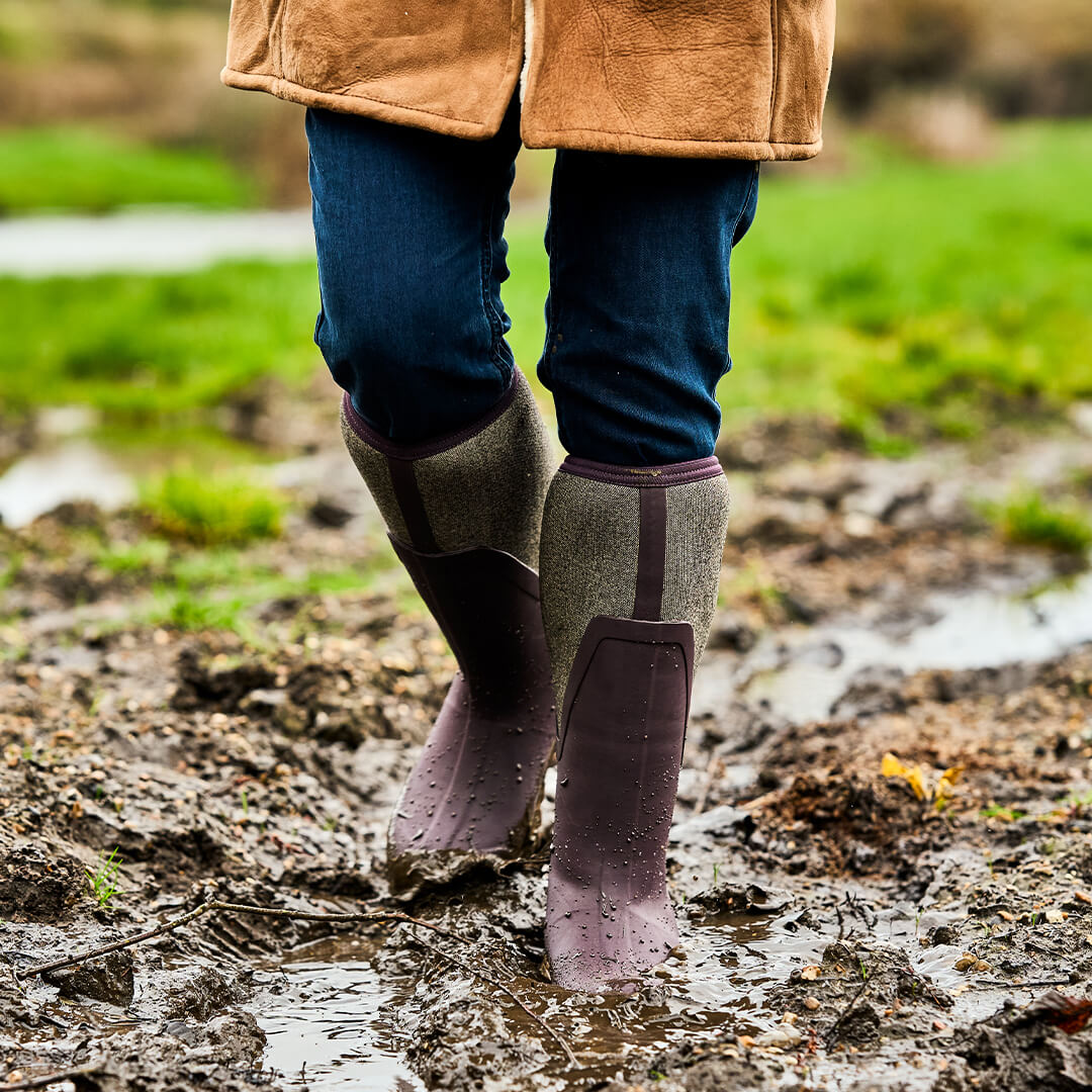 Eine Person in einem Paar Muck Boots Arctic Sport Tall Gummistiefel und einem Mantel, die über ein schlammiges Feld geht.